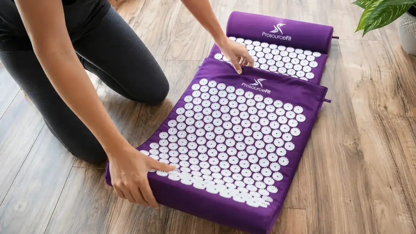 A close-up of hands unrolling a ProsourceFit acupressure mat with its plastic spike tiles and matching pillow on a wood floor