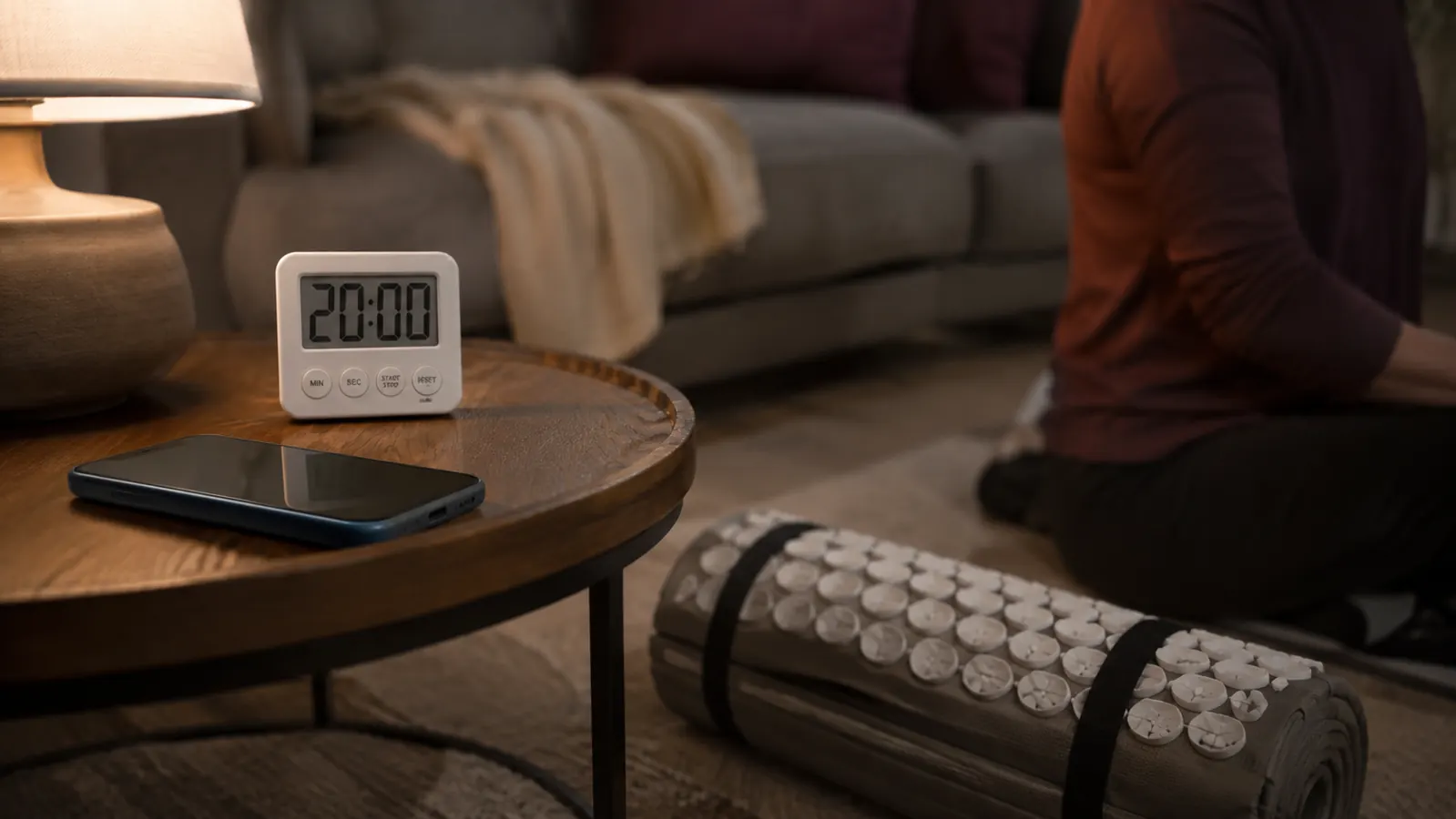 Phone placed face-down on a side table next to a rolled acupressure mat on the floor in a quiet evening room