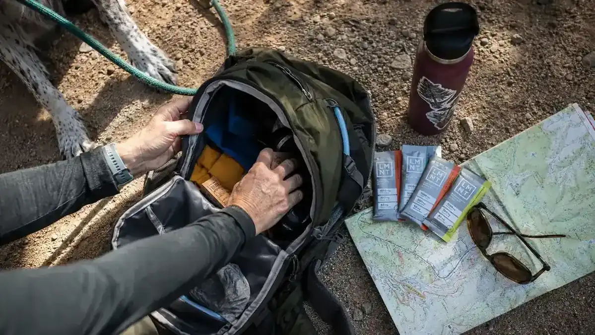 Open daypack on a trailhead bench with electrolyte packets tucked next to a water bottle and trail map