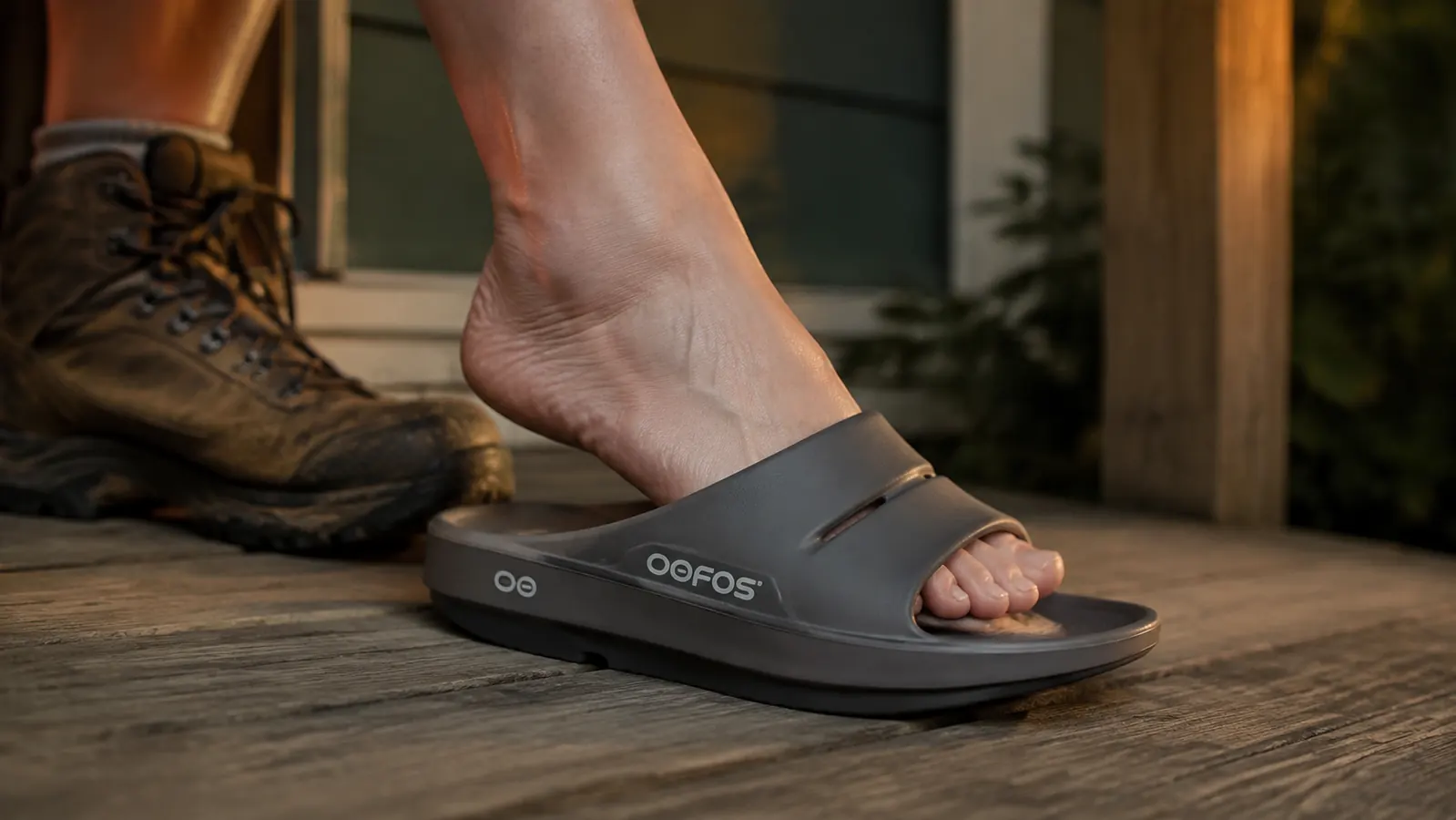 Close-up of a foot stepping out of a hiking boot directly into an OOFOS recovery slide on a wood porch