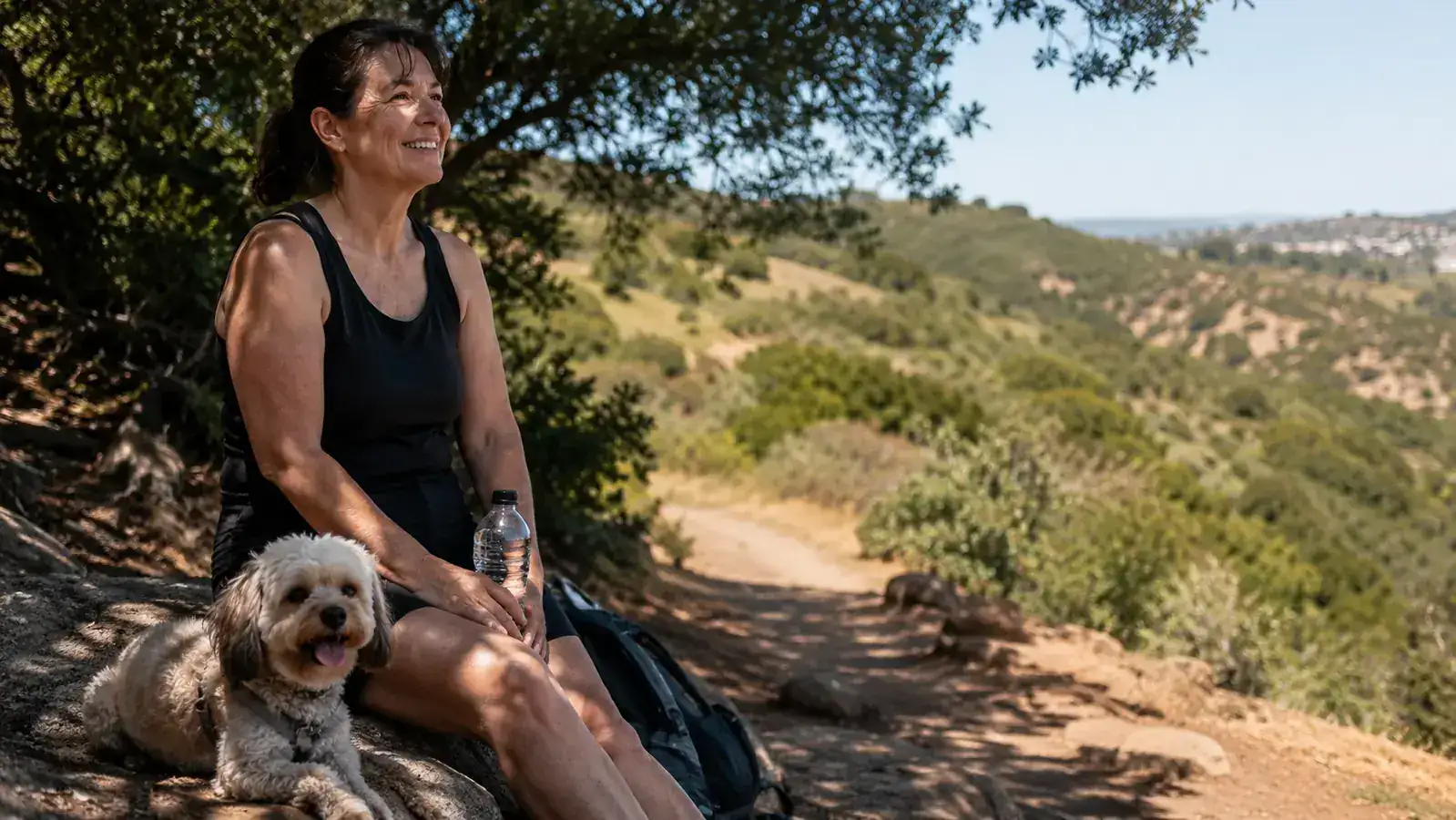 Woman and small dog sitting on a rock at an overlook mid-hike, water bottle in hand, wide trail view behind them