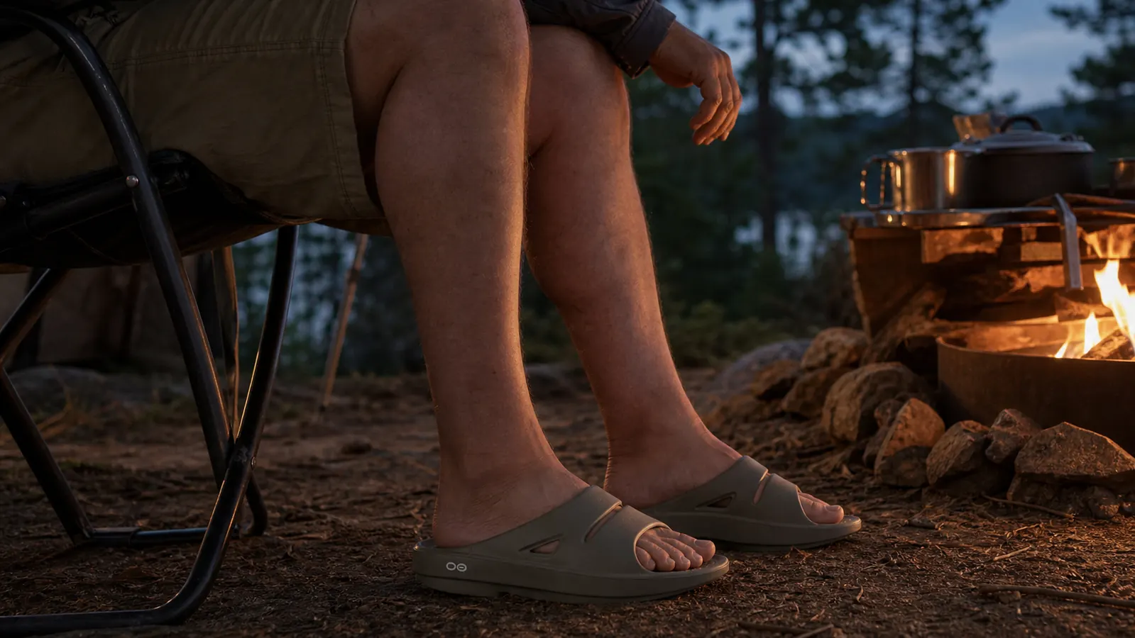 Bare feet in OOFOS recovery slides resting near a campfire with hiking boots set off to the side