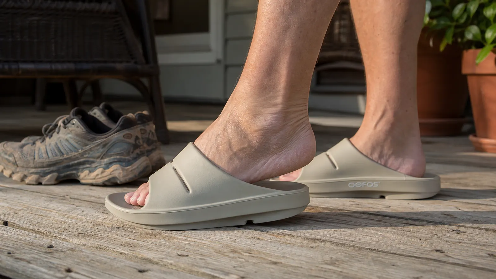 Bare feet stepping into OOFOS OOahh recovery slides next to a pair of trail sneakers on a cabin porch