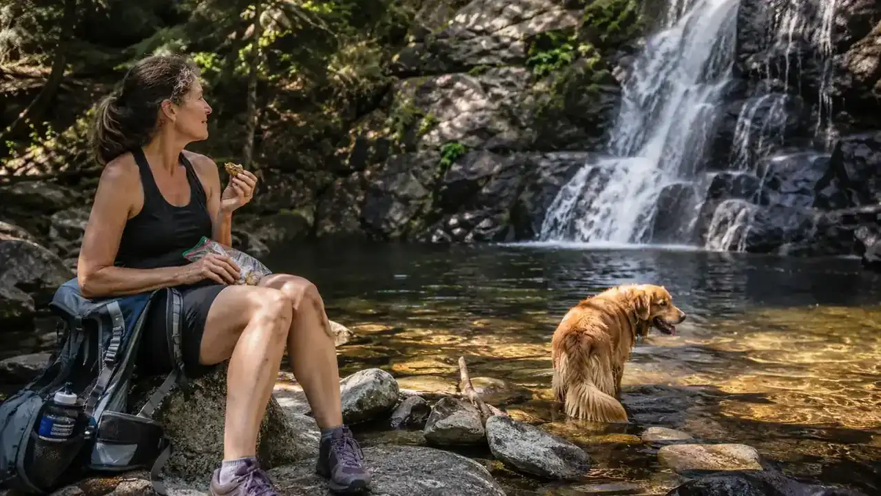 Woman and heeler mix dog standing near a waterfall pool at the end of a mountain trail