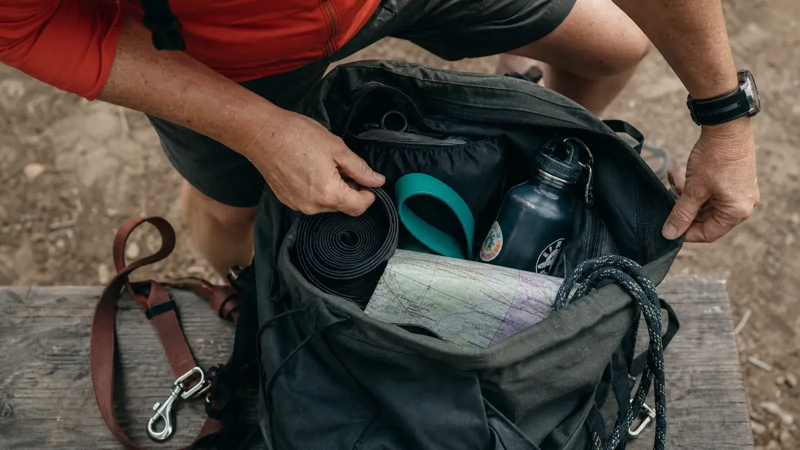 Daypack packed for a hike with a rolled stretching strap and a loop resistance band tucked beside a water bottle