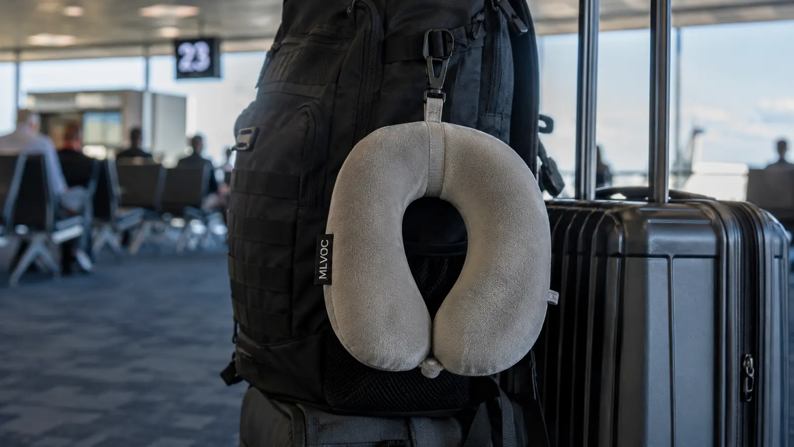 The MLVOC travel neck pillow clipped by its strap to the outside of a backpack next to a rolling suitcase at an airport gate