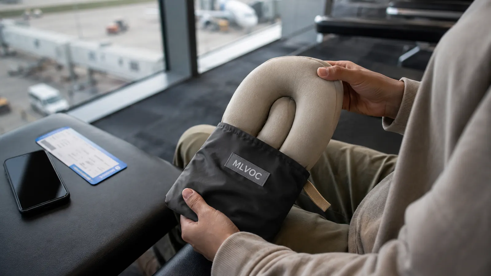 Hand holding the MLVOC travel neck pillow next to its compression pouch on an airport seat